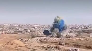 Explosion in Gaza releasing blue smoke, allegedly part of a gender reveal by Israeli soldiers, with urban ruins visible in the background.