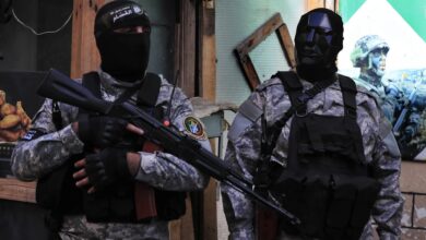 Two Hamas fighters in camouflage uniforms and black tactical gear stand armed with rifles in Lebanon. One wears a black face mask, the other a dark angular helmet mask. Urban setting with a military poster in the background.