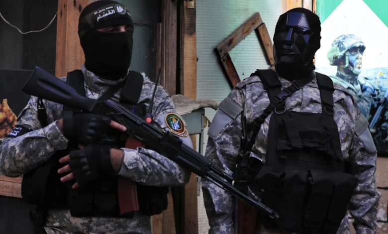 Two Hamas fighters in camouflage uniforms and black tactical gear stand armed with rifles in Lebanon. One wears a black face mask, the other a dark angular helmet mask. Urban setting with a military poster in the background.