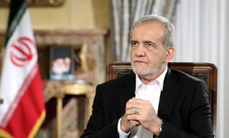 Iranian President Masoud Pezeshkian sits during a formal address, hands clasped, with the Iranian flag and a portrait in the background.