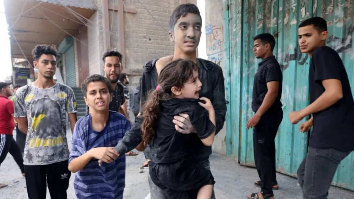 Dust-covered Palestinian children in Gaza walk through a bombed street after an Israeli airstrike, one carrying a terrified young girl to safety.