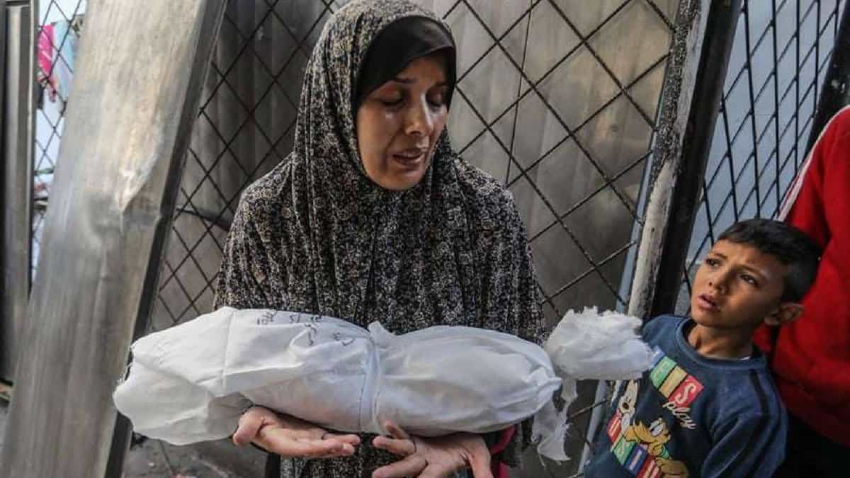 Grieving Palestinian mother holds her infant wrapped in a white shroud after Israeli airstrikes on Khan Younis, Gaza, as a young boy looks on in sorrow.