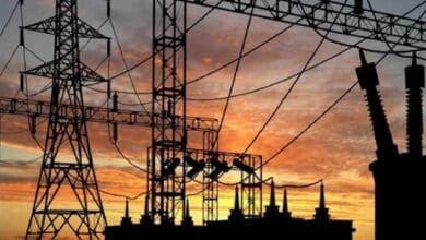 Silhouette of high-voltage transmission towers and power lines against a vibrant orange and pink sunset sky, with a power plant's cooling towers visible in the background for power cuts in Hyderabad story