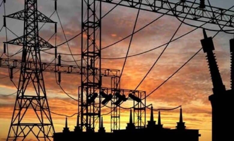 Silhouette of high-voltage transmission towers and power lines against a vibrant orange and pink sunset sky, with a power plant's cooling towers visible in the background for power cuts in Hyderabad story