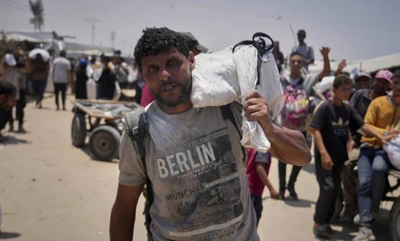The image of 43-year-old Omar al-Hobi from Rafah carrying a bag of food over his shoulder, wearing a "Berlin Munich" t-shirt, walks through a crowded area with tents and carts in the background in Gaza.
