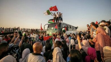 A large crowd of activists gathers around a bus as part of the Global March to Gaza. Several people stand on top of the bus waving Palestinian, Tunisian, and Algerian flags, while others cheer and film the scene with their phones. The sun sets in the background.