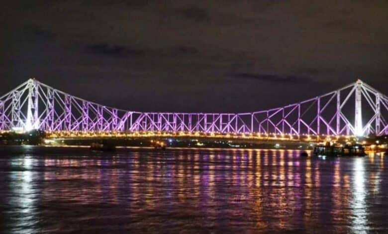 A night view of the Howrah Bridge in Kolkata, India, illuminated with purple lights and reflecting beautifully on the Hooghly River.