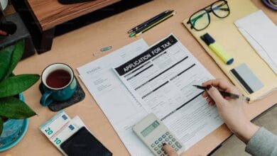 The image of a person’s hand holding a pen fills out a tax application form on a wooden desk, alongside an invoice, a calculator displaying £65, a cup of tea, euro banknotes, glasses, pencils, a highlighter, and a potted plant.