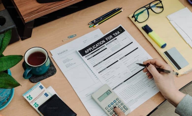 The image of a person’s hand holding a pen fills out a tax application form on a wooden desk, alongside an invoice, a calculator displaying £65, a cup of tea, euro banknotes, glasses, pencils, a highlighter, and a potted plant.