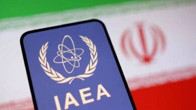 The image of a smartphone displaying the IAEA (International Atomic Energy Agency) logo is placed in front of a blurred Iranian flag, symbolising nuclear oversight and Iran's atomic programme.