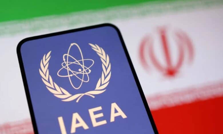 The image of a smartphone displaying the IAEA (International Atomic Energy Agency) logo is placed in front of a blurred Iranian flag, symbolising nuclear oversight and Iran's atomic programme.