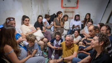 A group of men, women, and children sit closely together inside a public bomb shelter in Jerusalem. Some are using their phones, while others appear to be talking or waiting quietly, as they take cover during a missile alert.