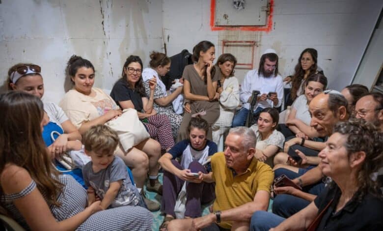 A group of men, women, and children sit closely together inside a public bomb shelter in Jerusalem. Some are using their phones, while others appear to be talking or waiting quietly, as they take cover during a missile alert.