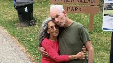 Weinstein, 70, and Haggai, 72, both Israeli-American hostages, embrace in front of a sign reading "Bayview Village Park." Weinstein, in a red shirt, has curly gray hair, and Haggai, in a green shirt, has white hair. A black trash bin and a notice about coyotes are in the background. They stand on a grassy area near a paved path.