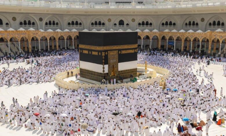 The image of a large crowd of pilgrims in white Ihram garments surrounds the Kaaba at the Grand Mosque in Makkah during daylight. The Kiswah covering the Kaaba is clearly visible, with the sacred door and golden embroidery in view.