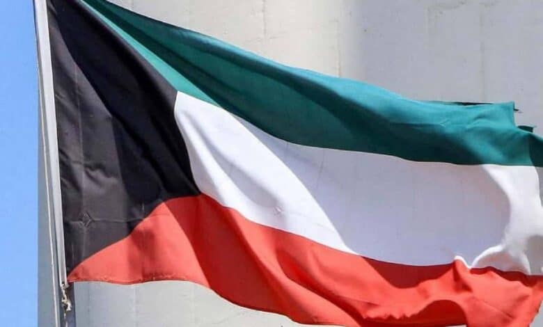 The national flag of Kuwait waving in the wind, showing horizontal stripes of green, white, and red with a black trapezoid on the hoist side, against a light-coloured background.