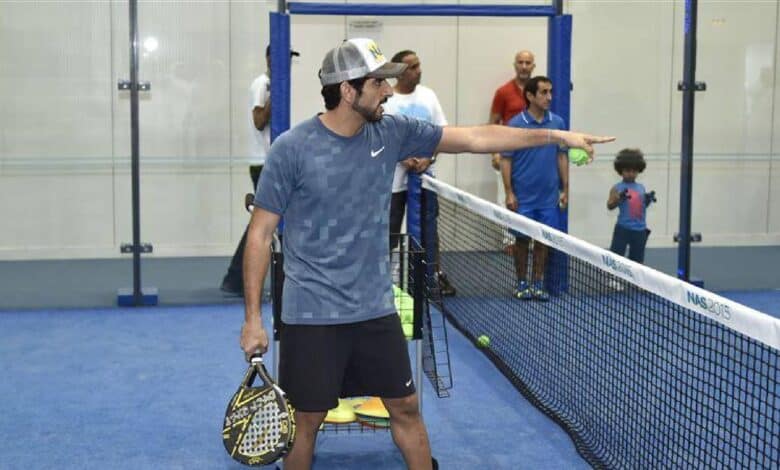Sheikh Hamdan bin Mohammed bin Rashid Al Maktoum, Crown Prince of Dubai, playing padel on an indoor court, holding a paddle, with other players and a child in the background.