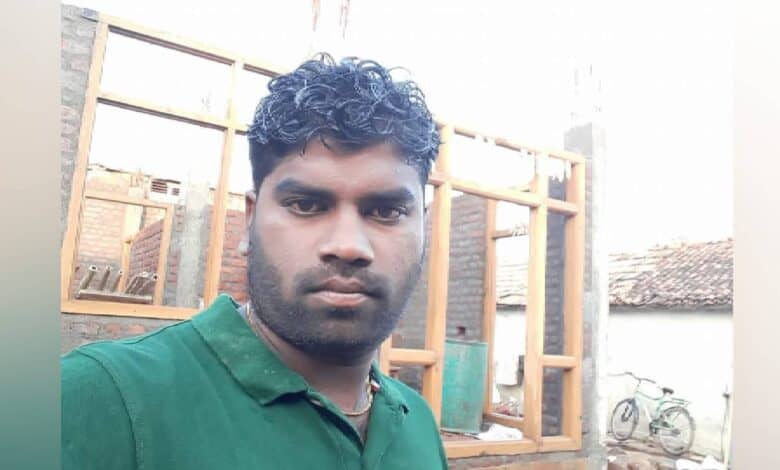The image of a man in a green shirt stands in front of a partially constructed brick building at a rural construction site.