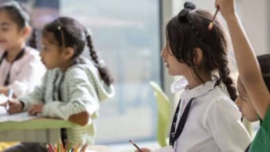 The image of a young children in a UAE classroom sit at desks, engaged in learning activities; one child raises a hand while others write or draw.