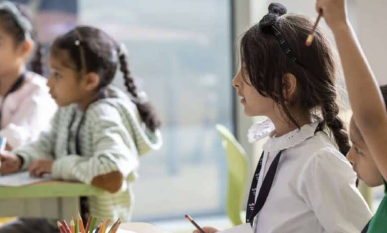 The image of a young children in a UAE classroom sit at desks, engaged in learning activities; one child raises a hand while others write or draw.