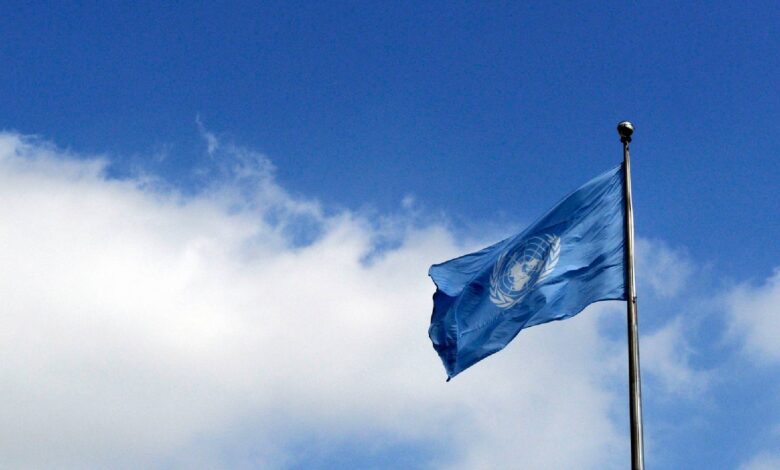 The image of United Nations flag waving against a bright blue sky with scattered clouds.