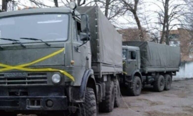 A line of olive‑drab military cargo trucks with canvas-covered beds, parked in a wooded yard — the lead vehicle marked with a prominent yellow “X.” Sources