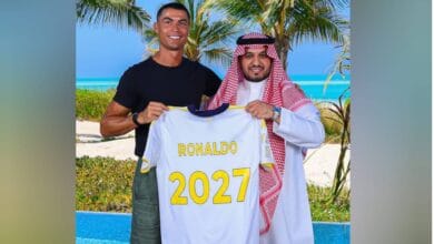 The image of Cristiano Ronaldo stands beside an Al Nassr official on a tropical beach, holding a football jersey with his name and "2027" printed on the back, celebrating his contract extension.