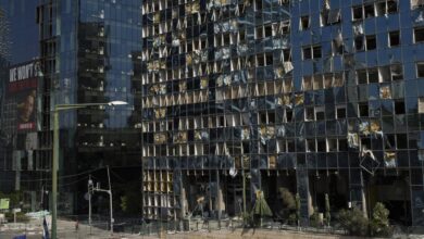 A damaged building with shattered windows and debris, located in Ramat Gan, Israel, following an Iranian missile strike on June 20, 2025. A member of the Israeli security forces walks past the structure, with a traffic light and a billboard reading 'WE WON'T' visible in the background.