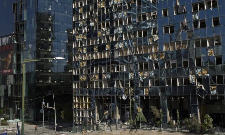 A damaged building with shattered windows and debris, located in Ramat Gan, Israel, following an Iranian missile strike on June 20, 2025. A member of the Israeli security forces walks past the structure, with a traffic light and a billboard reading 'WE WON'T' visible in the background.