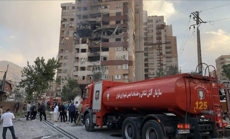 A damaged building in Tehran, Iran, with visible fire and structural collapse, surrounded by emergency response vehicles and personnel, set against a backdrop of trees and mountains, captured on June 13, 2025.