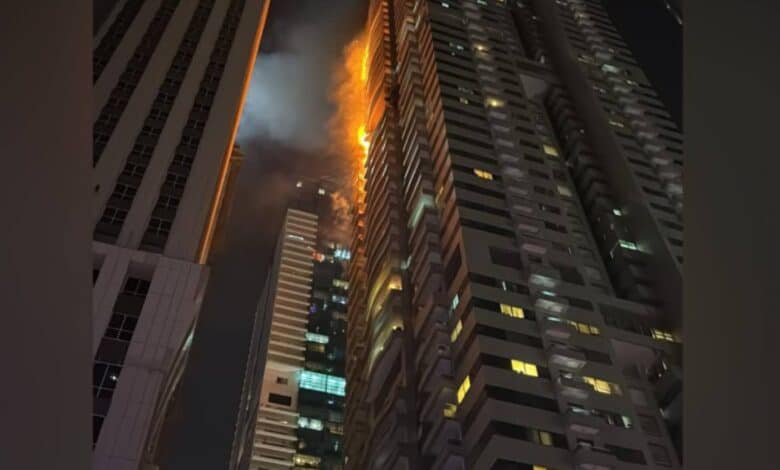 Flames and smoke rise from the upper floors of a high-rise residential tower at night in Dubai Marina, with neighbouring buildings visible in the foreground.