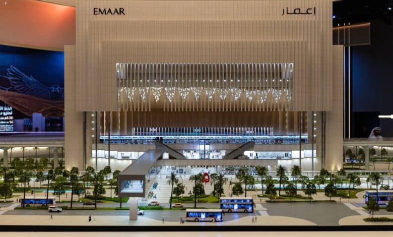 A detailed architectural model of the Emaar Properties Station on the Dubai Metro Blue Line, showcasing a futuristic and illuminated structure with pedestrian bridges, greenery, and buses in the foreground. The tall, modern design features vertical lighting and a grand facade.