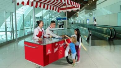 Two Emirates staff serve complimentary ice cream from a red and white striped cart to two young children at Dubai International Airport, with a modern terminal and travelators in the background.