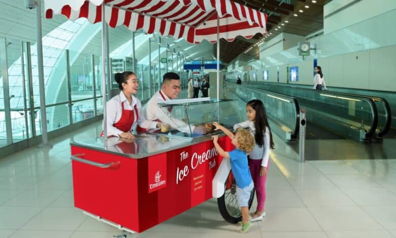 Two Emirates staff serve complimentary ice cream from a red and white striped cart to two young children at Dubai International Airport, with a modern terminal and travelators in the background.