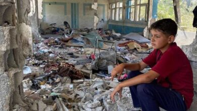 A Palestinian boy sits in the rubble of a destroyed classroom at a UNRWA school in Nuseirat, Gaza, surrounded by debris and torn belongings.