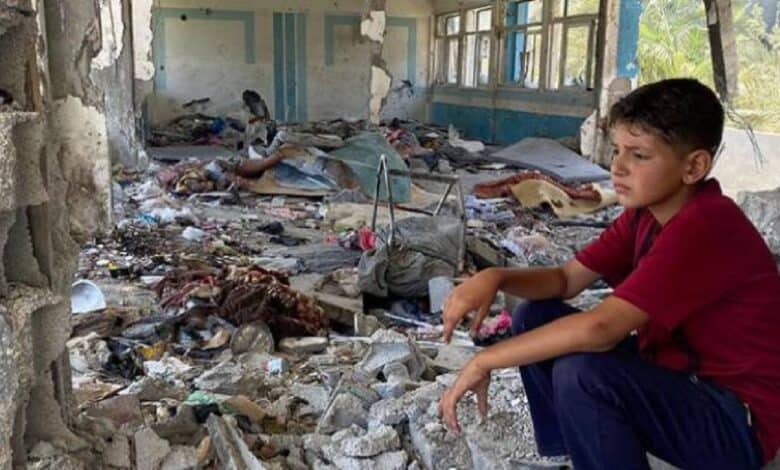 A Palestinian boy sits in the rubble of a destroyed classroom at a UNRWA school in Nuseirat, Gaza, surrounded by debris and torn belongings.