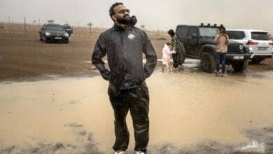 Muhammed Sajjad, known locally as “The Weatherman”, stands in a puddle wearing a black raincoat and flip-flops during rain in the Sharjah desert, with off-road vehicles and other participants in the background.