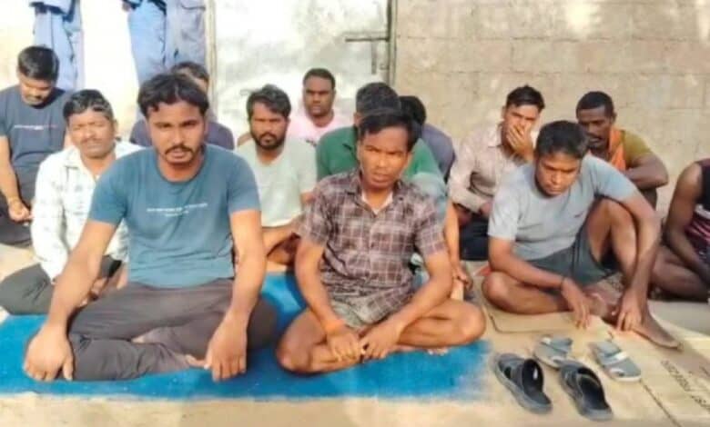 The image of a group of Indian migrant workers sit on mats and cardboard sheets against a wall, some looking directly at the camera while others appear visibly distressed.