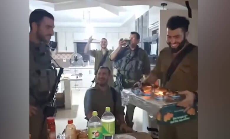 The image of a Israeli soldiers in uniform celebrate a birthday inside a Palestinian home, with one holding a cake and others clapping and filming the scene.