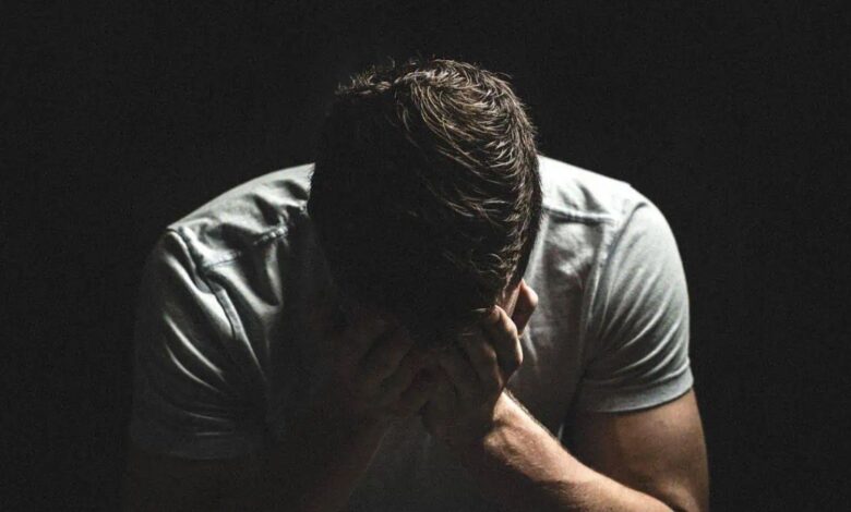 A shadowy image of a man with his head bowed and hands covering his face, wearing a gray shirt. The dark background emphasizes a mood of despair or deep thought, creating a somber and introspective atmosphere.