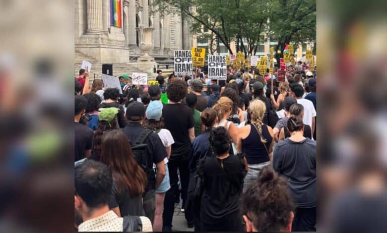 Large crowd gathered outside the New York Public Library during an anti-war rally, with placards reading “HANDS OFF IRAN” and “END ALL U.S. AID TO ISRAEL” visible above the crowd. Trees and skyscrapers frame the scene.