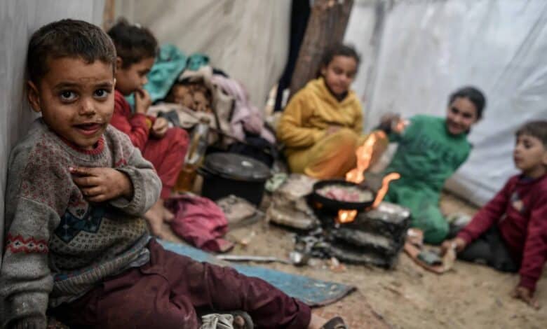 A group of Palestinian children in worn clothing sit inside a makeshift tent, with one child in the foreground looking at the camera, their face and jumper smeared with dirt. In the background, children gather around a small open fire cooking food in a pan.