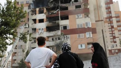 The image of a three people stand looking at a damaged residential building in Tehran, Iran, with blackened windows and visible structural destruction caused by an airstrike. A firefighter’s ladder reaches up to the upper floors.