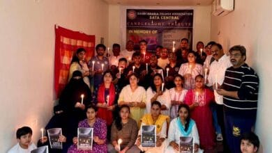 A group of Telugu NRIs in Riyadh, including men, women, and children, hold candles and condolence posters during a candlelight tribute for Air India AI-171 crash victims. A banner in the background reads "SATA Central Candlelight Tribute".