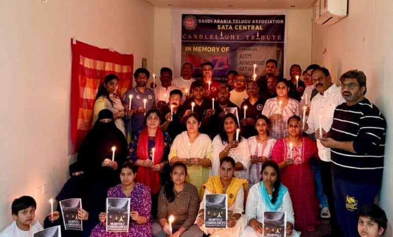 A group of Telugu NRIs in Riyadh, including men, women, and children, hold candles and condolence posters during a candlelight tribute for Air India AI-171 crash victims. A banner in the background reads "SATA Central Candlelight Tribute".