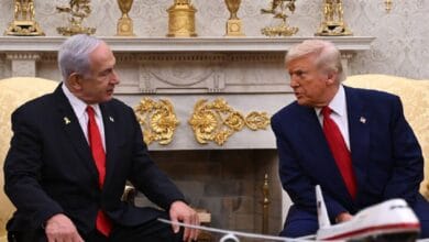US President Donald Trump (right) and Israeli Prime Minister Benjamin Netanyahu (left) sit facing each other in a formal setting, likely the Oval Office. Both are wearing dark suits with red ties. A model airplane is placed on the table between them. Behind them is an ornate fireplace decorated with gold vases and intricate embellishments, highlighting the room's grandeur.