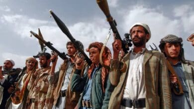 The image of a group of armed Houthi fighters in Yemen holding rocket-propelled grenades and rifles, standing in a line under a cloudy sky.