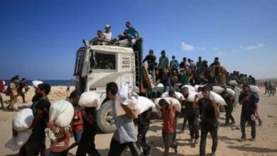 Palestinians carry sacks of aid as others ride atop a truck in Beit Lahia, northern Gaza, amid ongoing humanitarian crisis.
