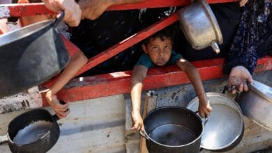 A young child with a somber expression peers out from a weathered red-framed structure in Gaza, clutching a metal pot. Surrounding hands of adults, also holding pots and pans, are visible.