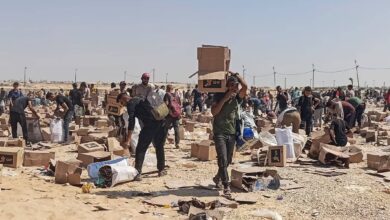 The image of people gather at a Gaza aid distribution site, collecting boxes and supplies scattered across a dusty, open area under the sun.